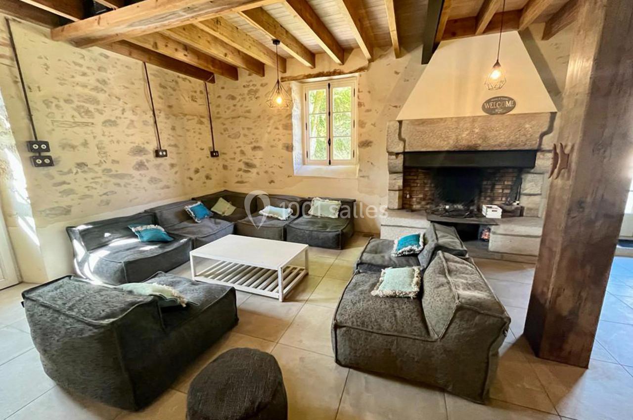 Salon chaleureux avec murs en pierre, poutres apparentes, cheminée, canapés gris et table basse blanche.