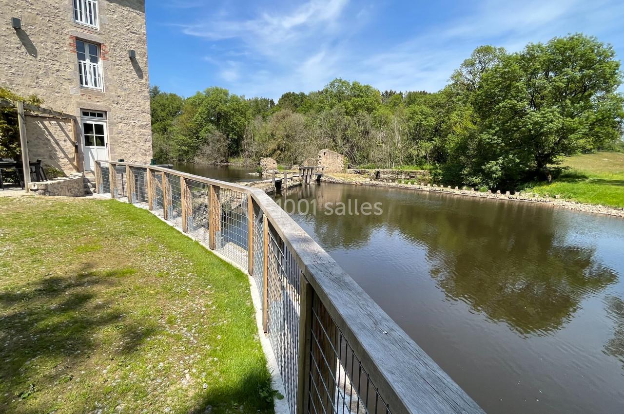 Vue d'un moulin en pierre près d'une rivière bordée d'arbres, avec une passerelle en bois au premier plan.