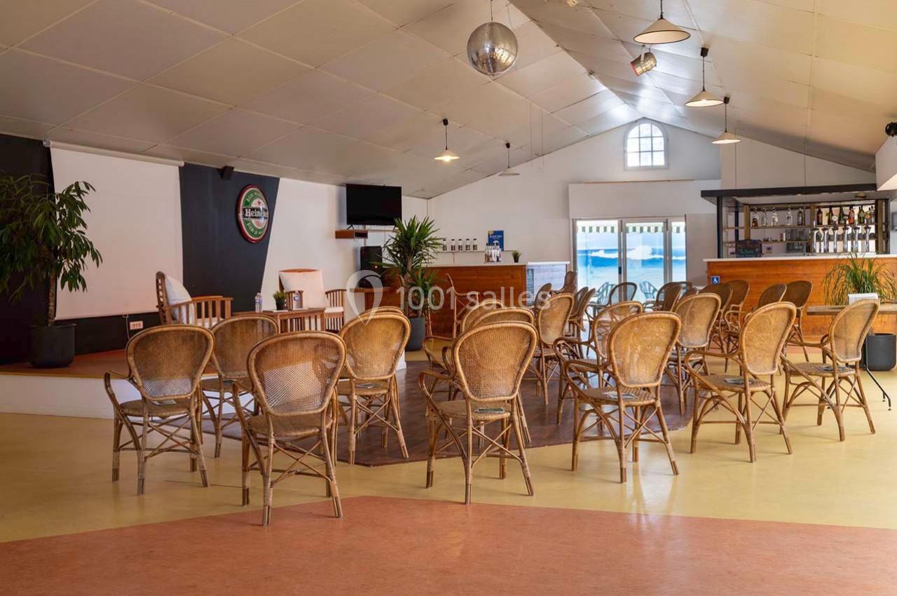 Salle lumineuse avec des chaises en rotin disposées face à une petite scène et un bar en arrière-plan.