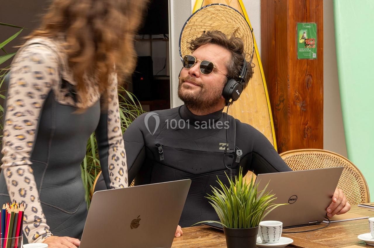 Un homme en combinaison de surf avec un casque audio travaille sur un ordinateur portable, une femme à ses côtés.