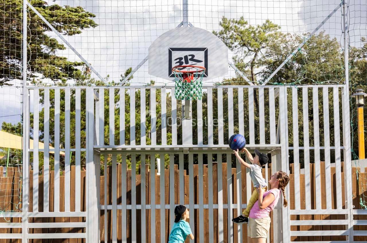 Une femme aide un enfant à marquer un panier de basket, sous le regard d'un autre enfant, dans un espace extérieur clôturé.