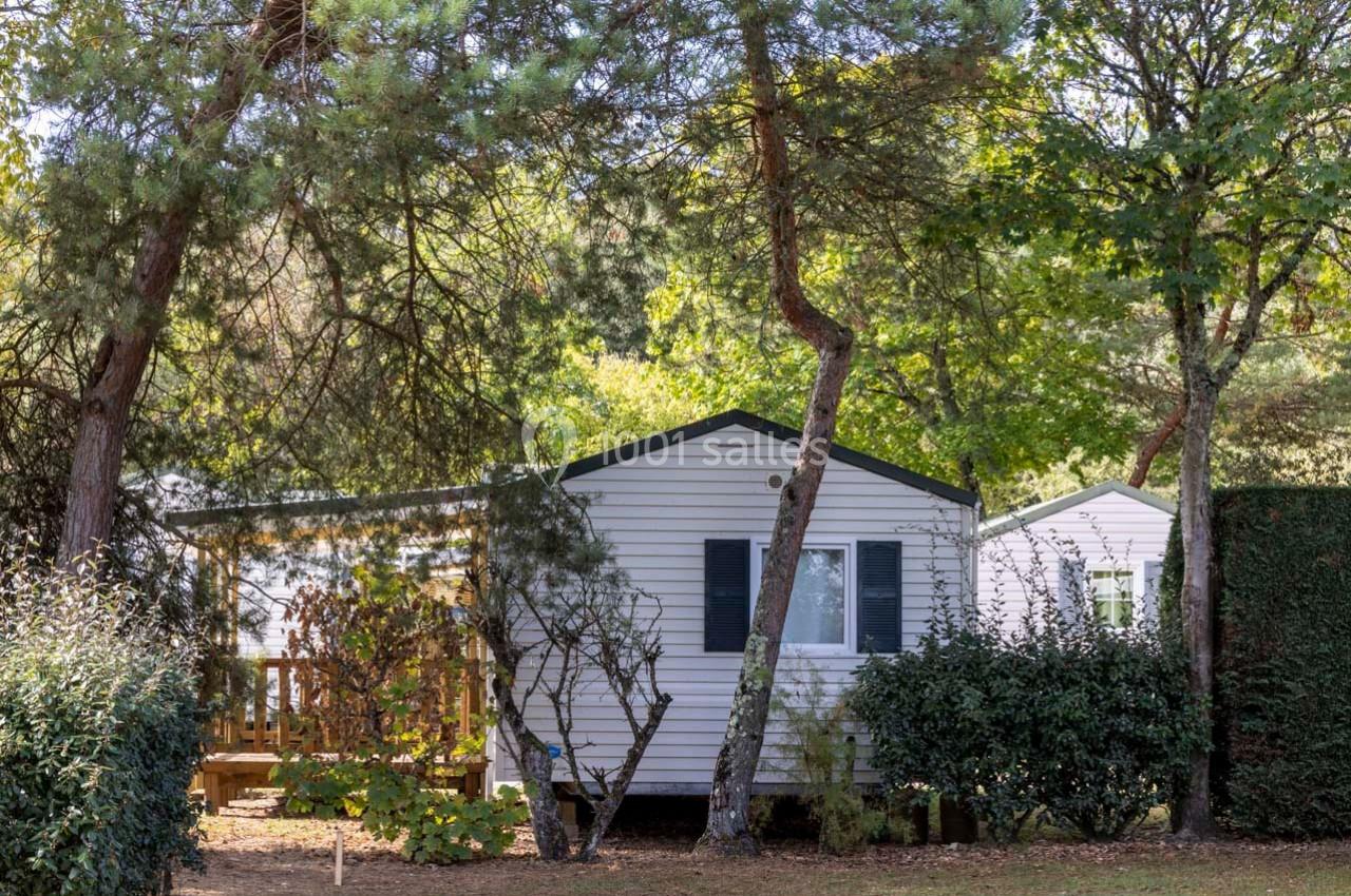 Mobil-home blanc entouré de végétation, avec une terrasse en bois partiellement visible sous des arbres.