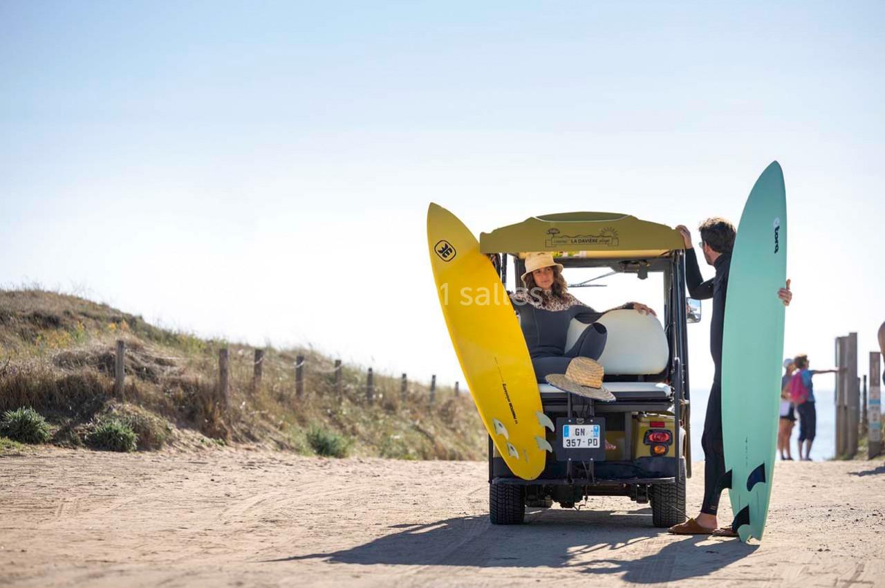 Deux personnes chargent des planches de surf sur une voiturette de golf stationnée sur un chemin sablonneux.