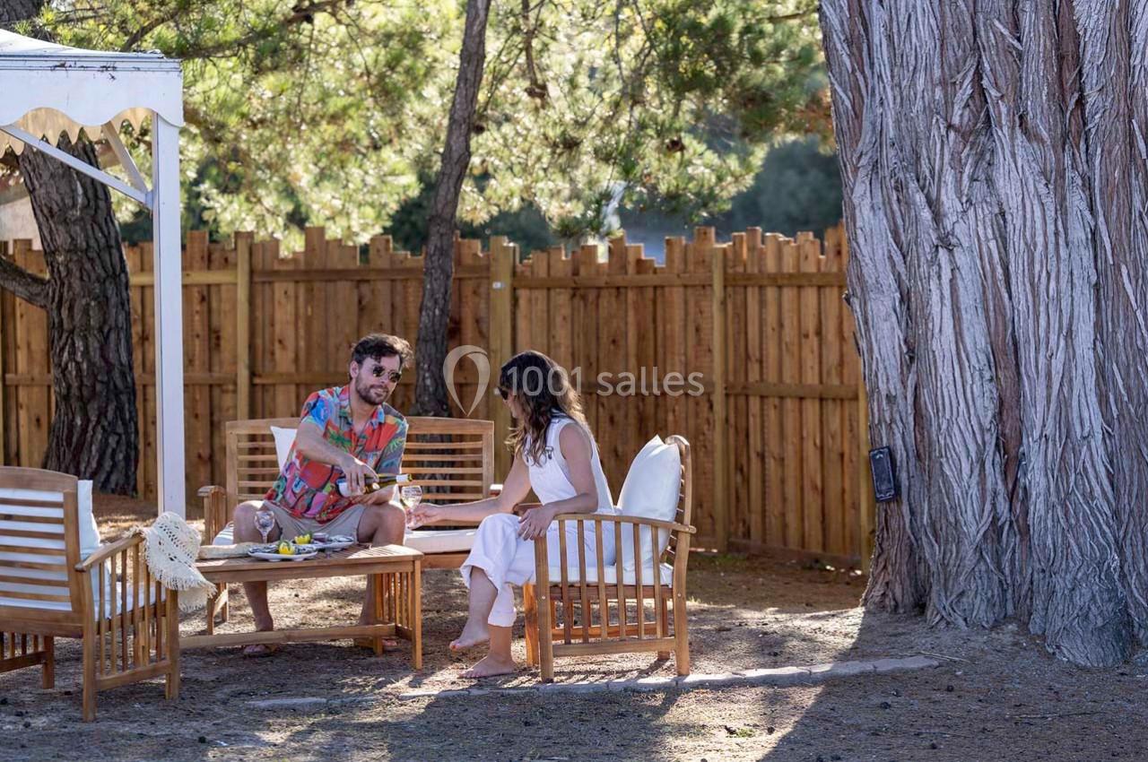 Un homme et une femme discutent assis sur des fauteuils en bois dans un jardin ombragé près d'un grand arbre.