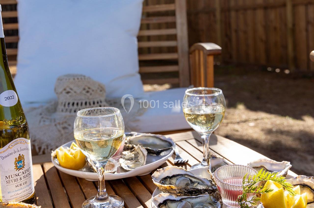 Table en bois avec assiettes d'huîtres, verres de vin blanc, citron et bouteille, dans un jardin ensoleillé avec fauteuil.