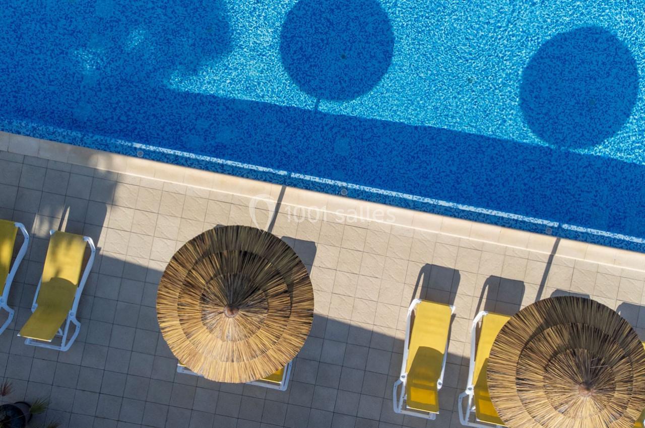 Vue aérienne de chaises longues jaunes et parasols en paille alignés près d'une piscine bleue.