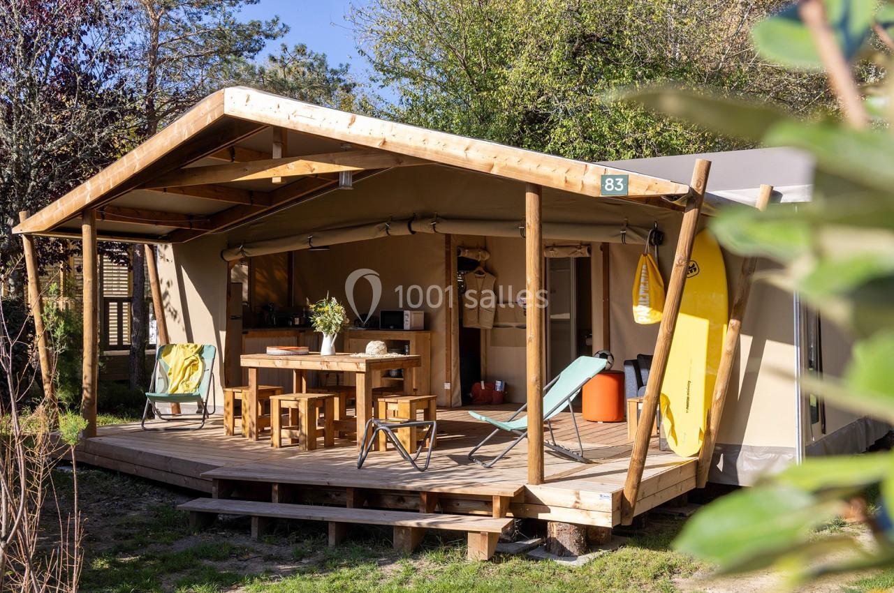 Tente lodge en bois avec terrasse aménagée, mobilier extérieur et équipements, entourée de verdure.