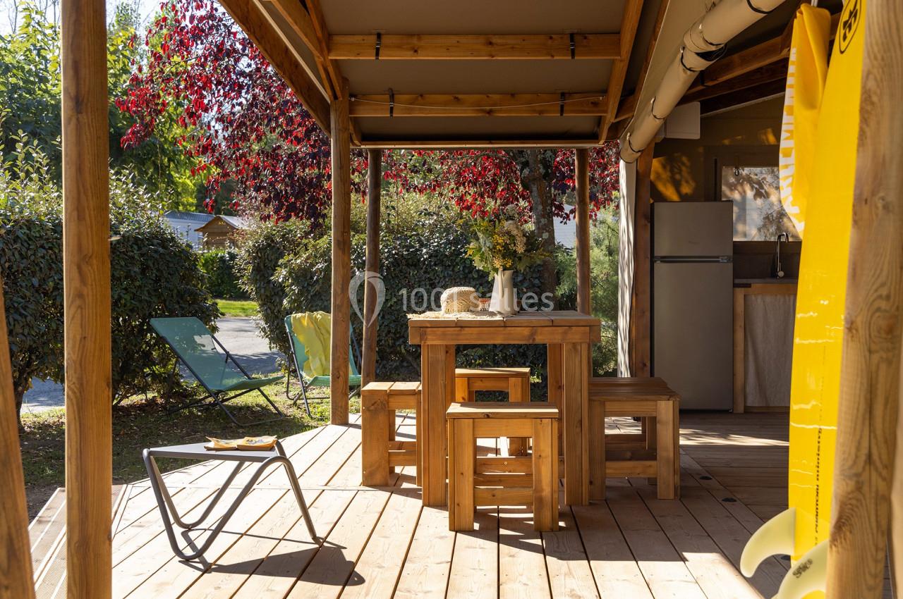 Terrasse en bois avec table et bancs, entourée de verdure, équipée de chaises longues et d'un coin cuisine en arrière-plan.