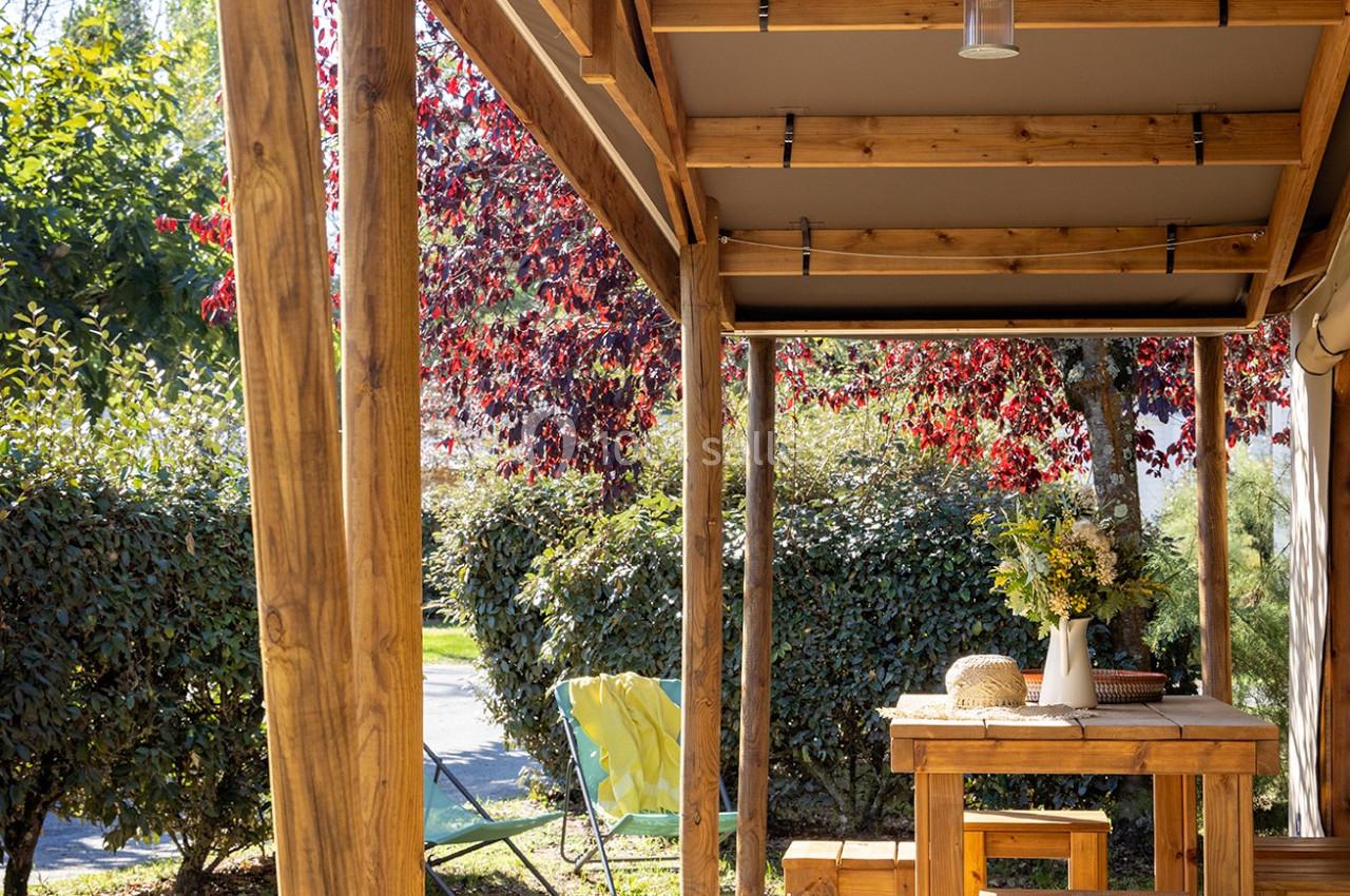 Terrasse en bois avec table, chaises et lanterne suspendue, entourée de végétation et éclairée par le soleil.
