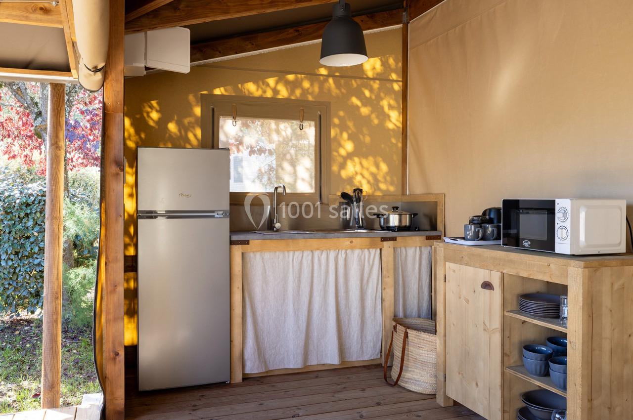 Cuisine extérieure en bois sous une tente, équipée d'un réfrigérateur, d'un évier, d'un micro-ondes et d'ustensiles.