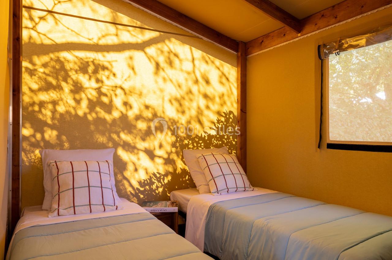 Deux lits simples avec des draps bleus dans une tente éclairée par la lumière du soleil filtrée à travers des arbres.