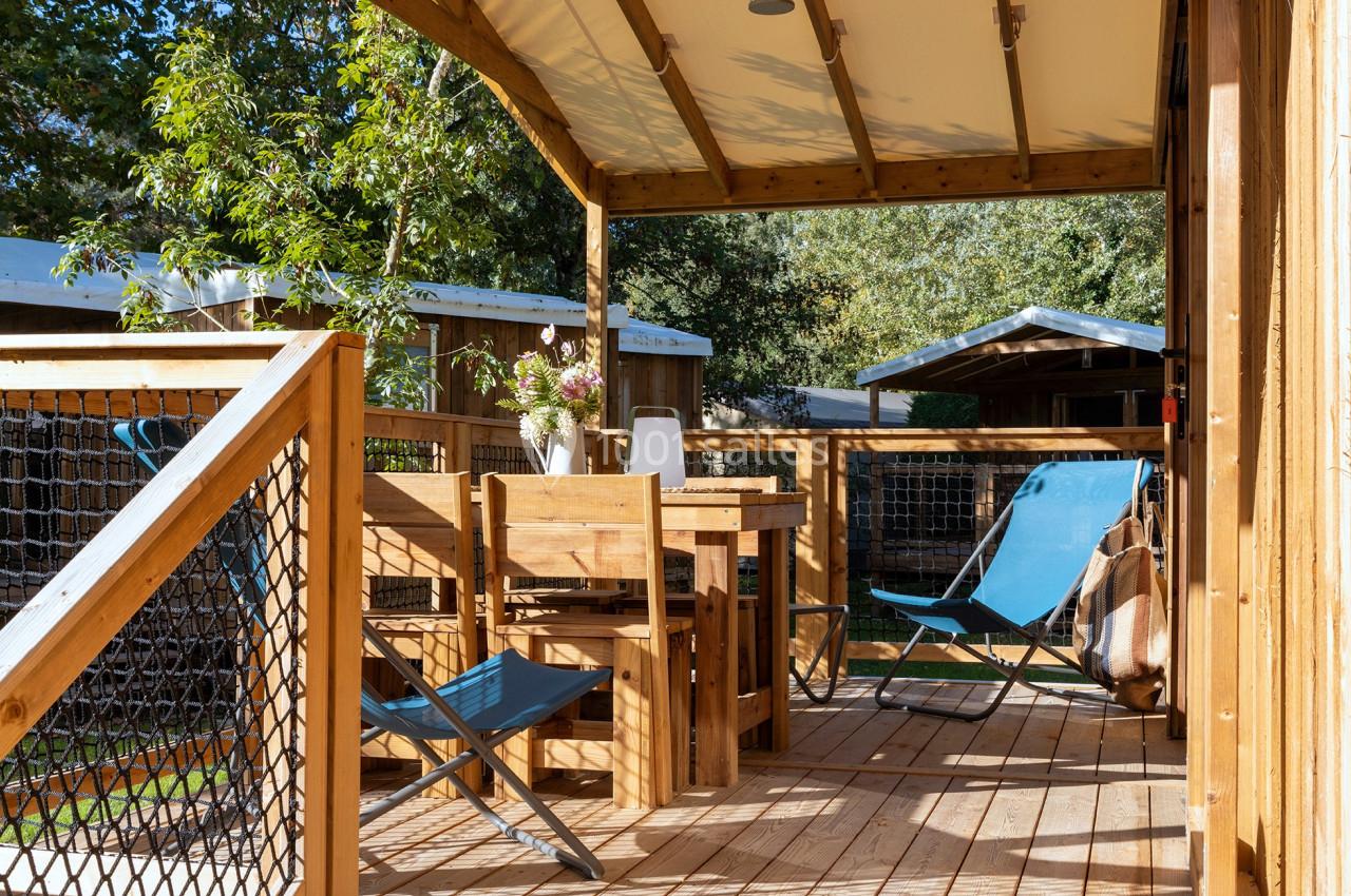 Terrasse en bois avec table, chaises, transats bleus et plantes, entourée de verdure et de structures en bois.