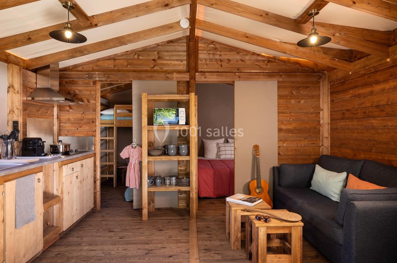 Intérieur d'un chalet en bois avec cuisine équipée, coin salon, lit, étagères et guitare posée près d'une table basse.
