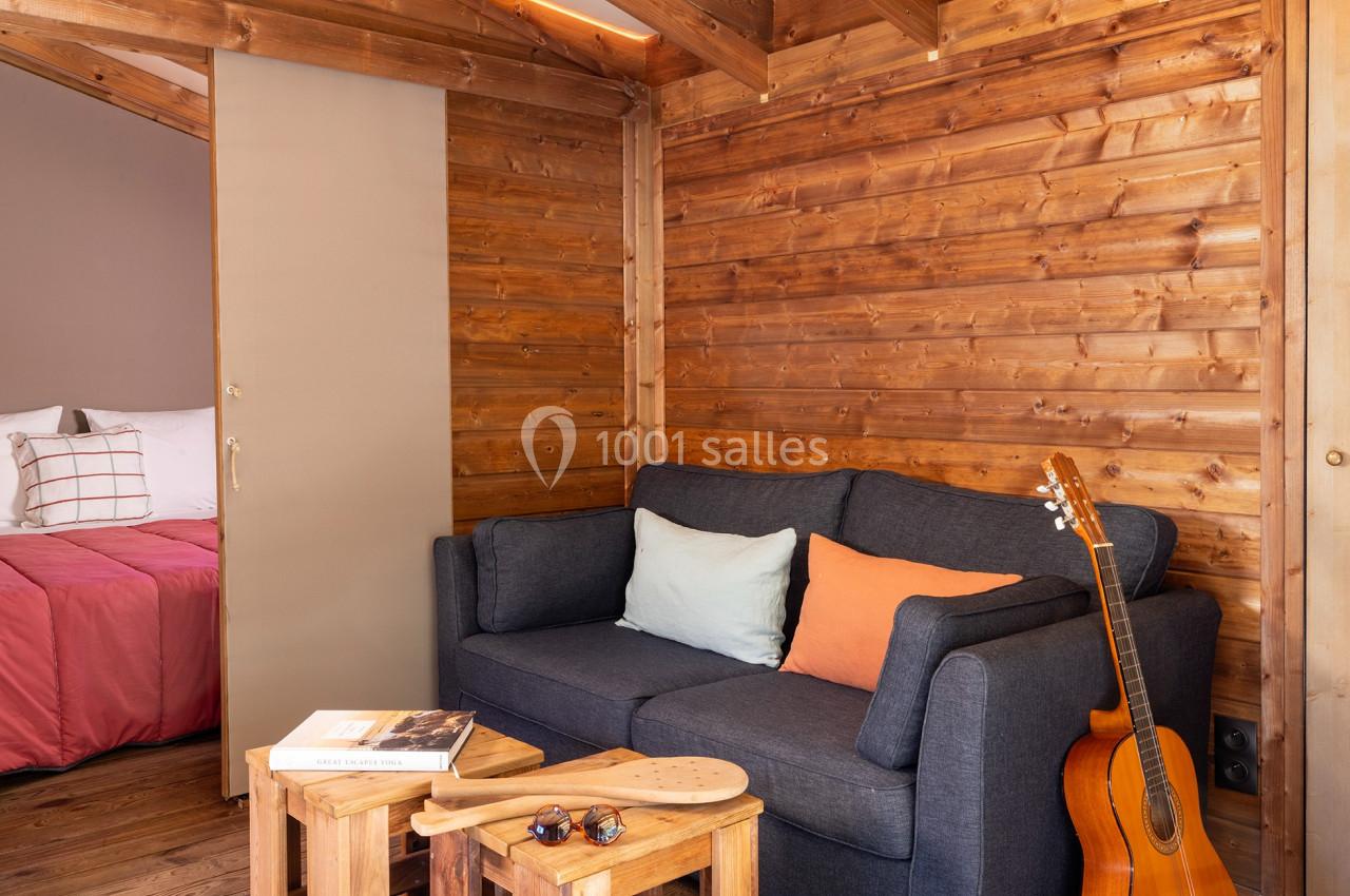 Salon en bois avec canapé gris, coussins colorés, guitare posée à côté et chambre visible en arrière-plan.