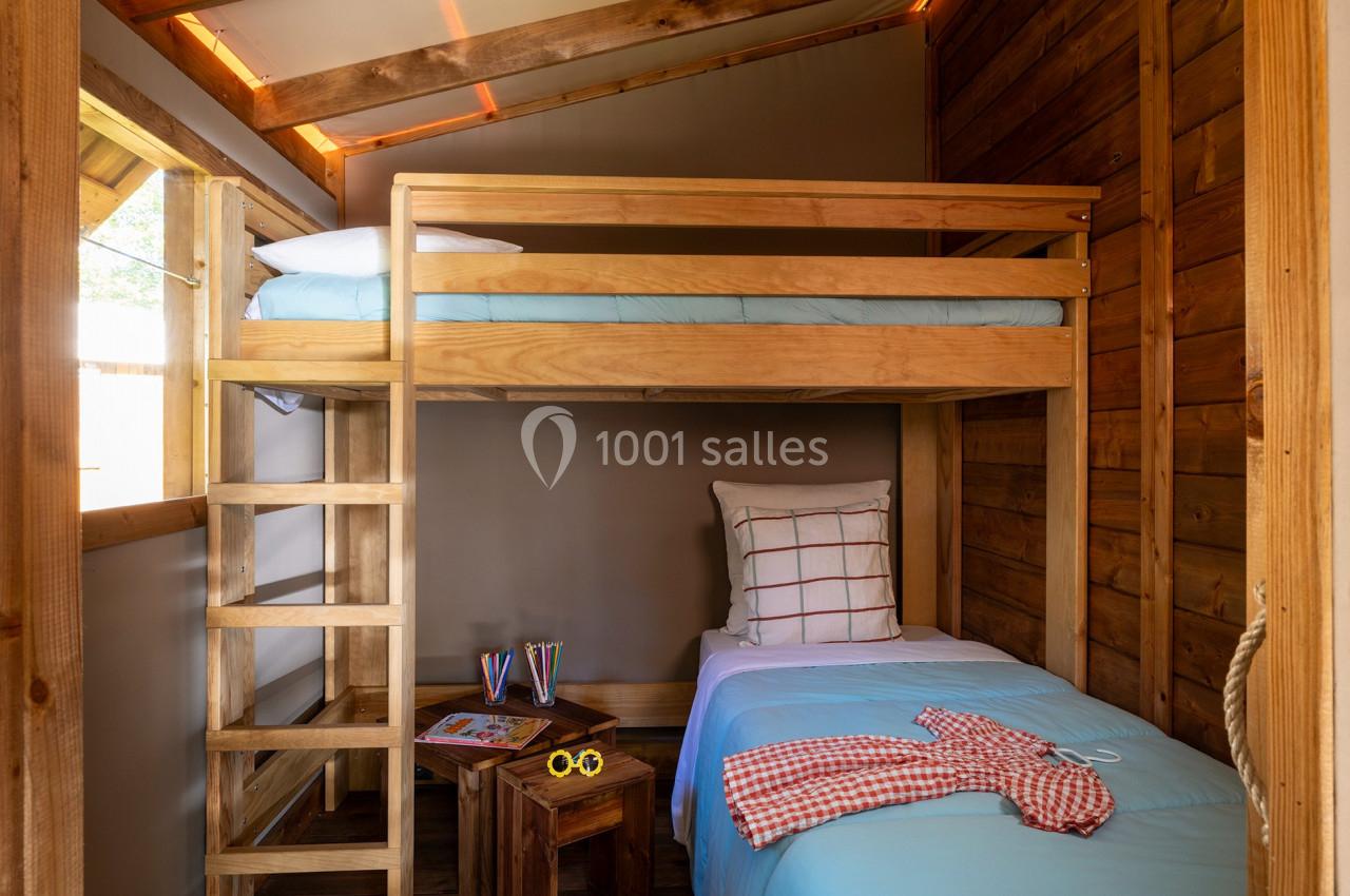 Lit superposé en bois dans une chambre rustique avec un lit simple en dessous, table de chevet et lumière naturelle.