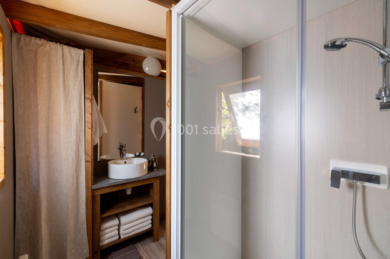 Salle de bain avec douche vitrée, lavabo sur meuble en bois, miroir, étagères avec serviettes et fenêtre lumineuse.