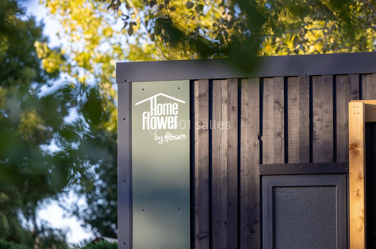 Façade en bois sombre d'un bâtiment avec un logo ’Home Flower by Bioser’, entourée de végétation et éclairée par le soleil.