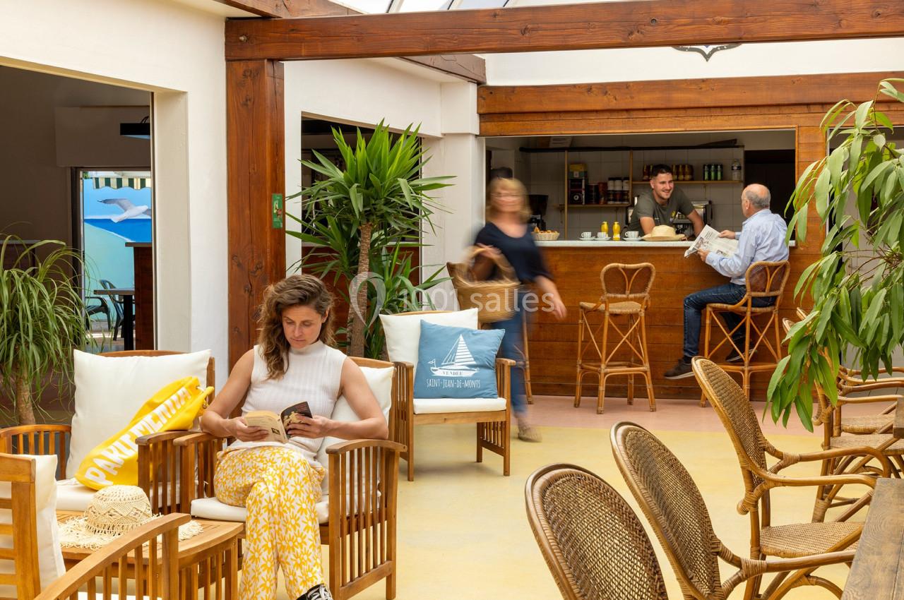 Une femme lit dans un salon lumineux avec des plantes, tandis que des personnes discutent au comptoir en arrière-plan.