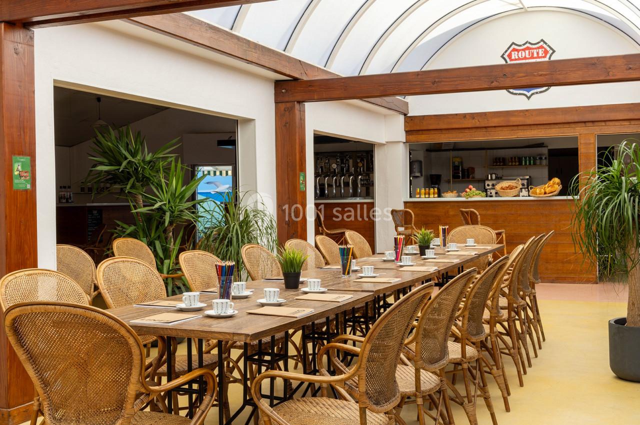 Salle lumineuse avec une grande table en bois, chaises en rotin, plantes vertes et un comptoir en arrière-plan.
