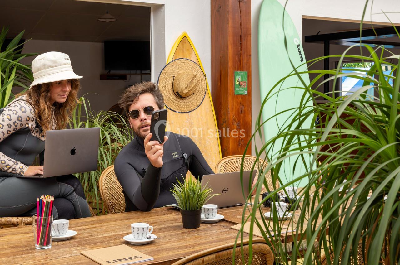 Deux personnes en tenue de surf travaillent sur des ordinateurs portables à une table entourée de plantes et de planches de…