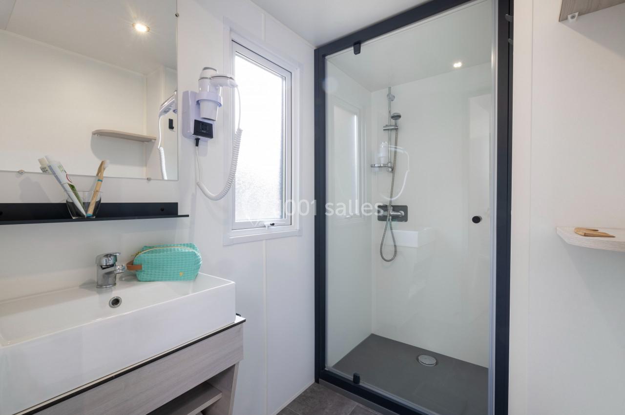 Salle de bain moderne avec douche vitrée, lavabo avec miroir, sèche-cheveux mural et fenêtre apportant de la lumière…