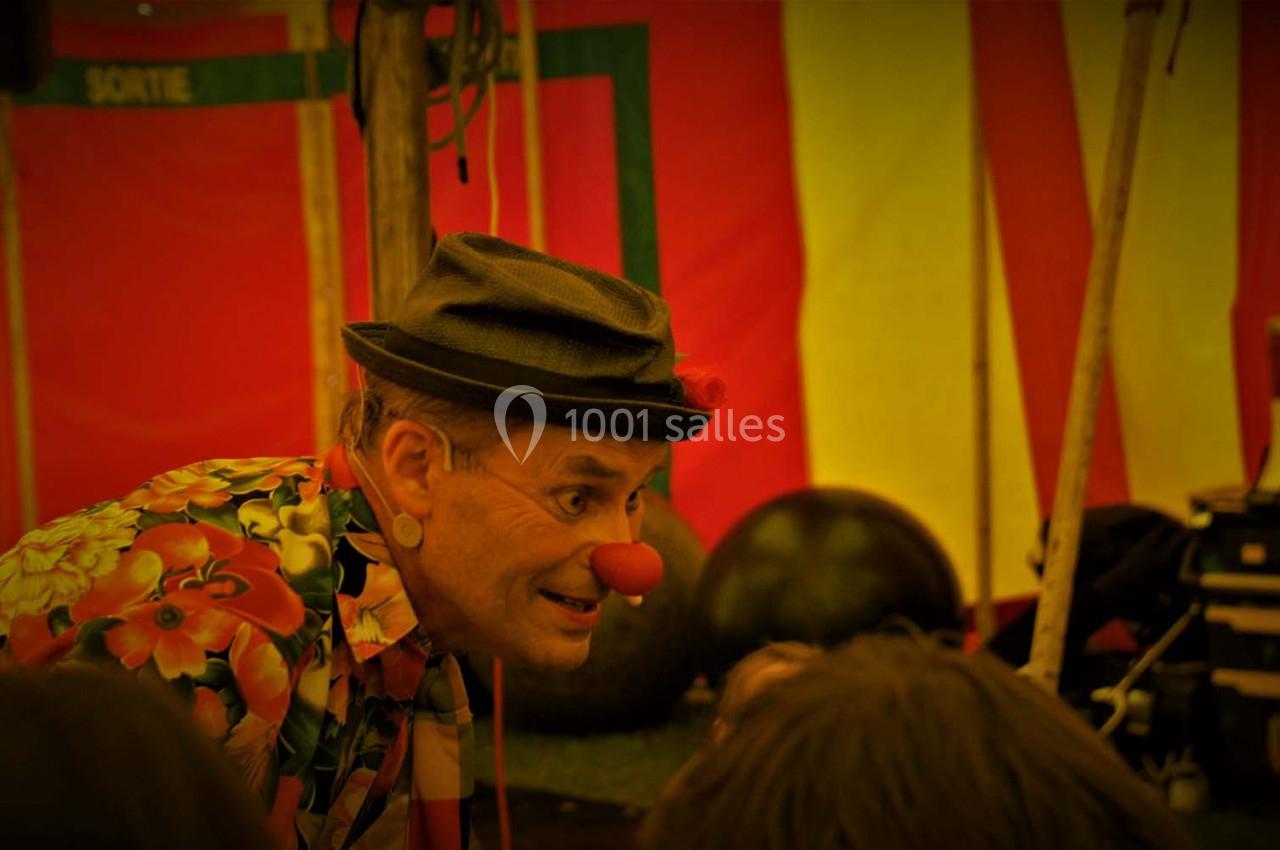 Un clown portant un chapeau et une chemise colorée interagit avec des enfants sous un chapiteau.