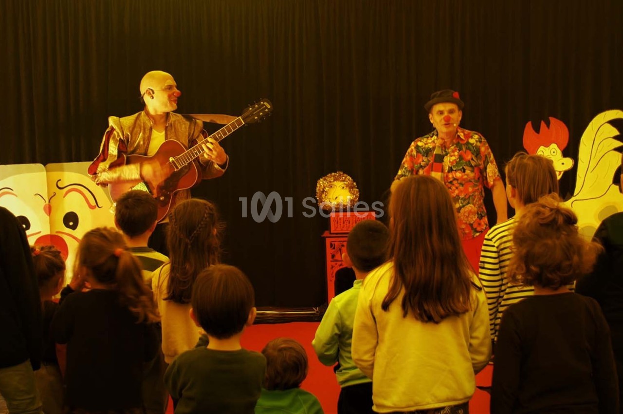 Des enfants assistent à un spectacle avec deux artistes, l’un jouant de la guitare, dans une salle décorée de motifs colorés.