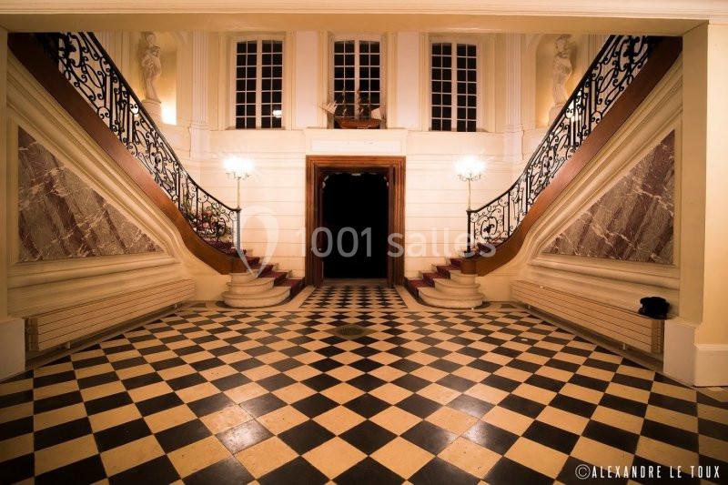 Hall d'entrée avec sol en damier, double escalier symétrique en marbre et balustrades en fer forgé.