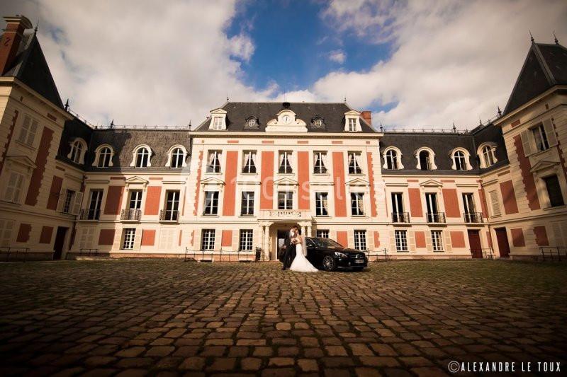 Un couple en tenue de mariage pose devant un château aux façades claires sous un ciel partiellement nuageux.
