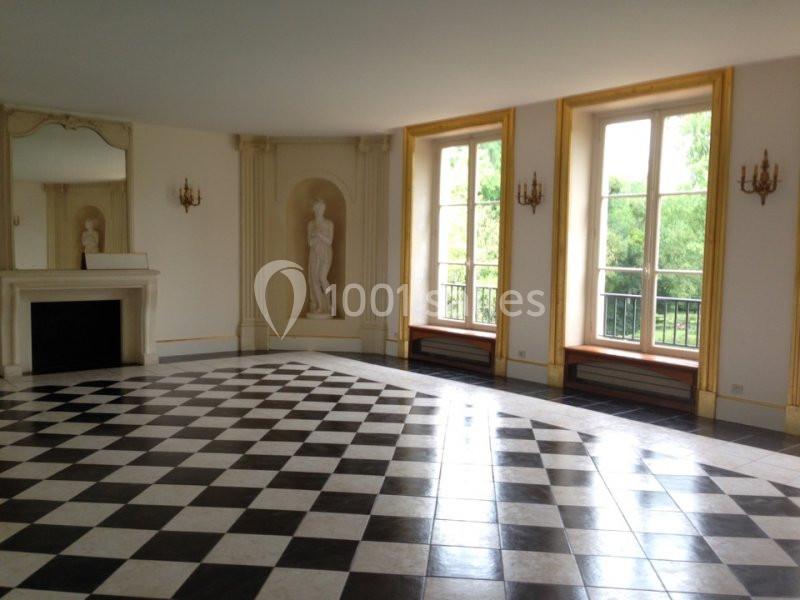 Salle lumineuse avec sol en damier noir et blanc, cheminée, statues décoratives et grandes fenêtres donnant sur un jardin.