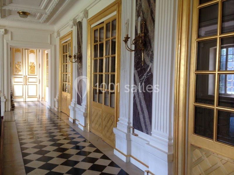 Couloir élégant avec sol en damier, portes vitrées dorées, murs ornés de marbre et appliques murales.