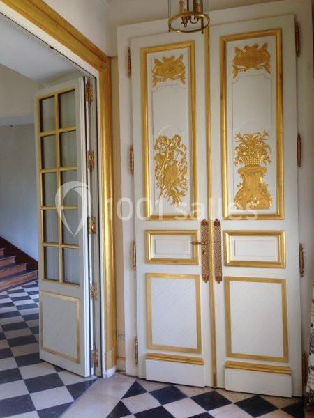 Porte double en bois blanc ornée de motifs dorés, dans une pièce avec carrelage noir et blanc et escalier en arrière-plan.