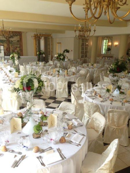 Salle de réception élégante avec tables rondes dressées, nappes blanches, chaises couvertes et décorations florales.