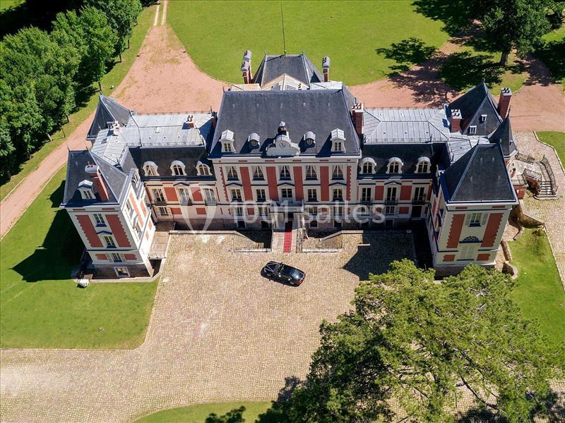 Vue aérienne d'un château avec façade symétrique, entouré de pelouses et d'une allée pavée où est garée une voiture.