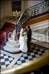 Un couple de mariés s'embrasse au pied d'un escalier élégant avec une rampe en fer forgé et un sol en damier.