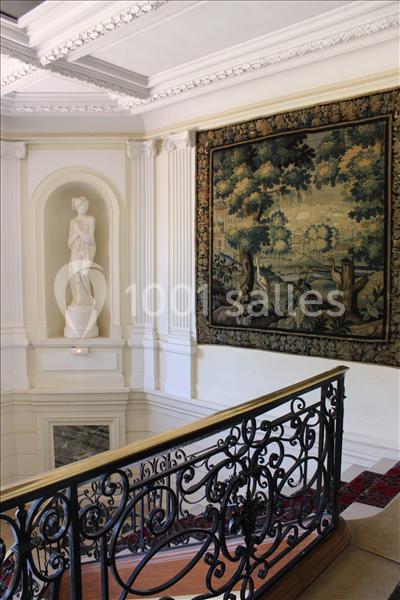 Escalier avec rampe en fer forgé, statue classique sur un socle et tapisserie murale représentant un paysage boisé.