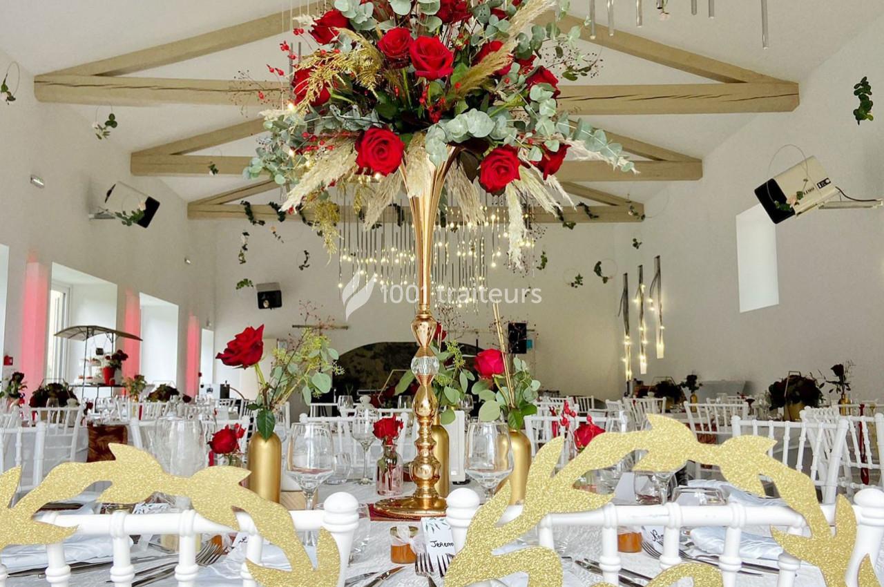 Centre de table floral avec roses rouges et feuillage, dans une salle de réception décorée avec des lumières suspendues.