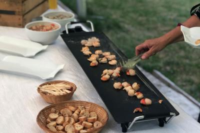 Personne cuisant des fruits de mer sur une plancha, avec des paniers de pain et des assiettes vides à proximité.