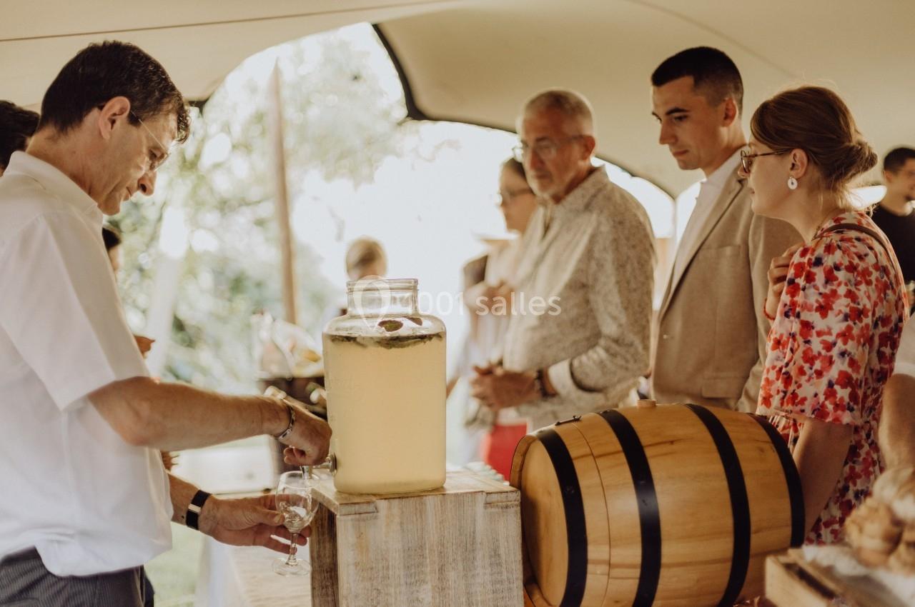 Un homme sert une boisson dans un verre à partir d'un grand bocal, avec des personnes discutant en arrière-plan.