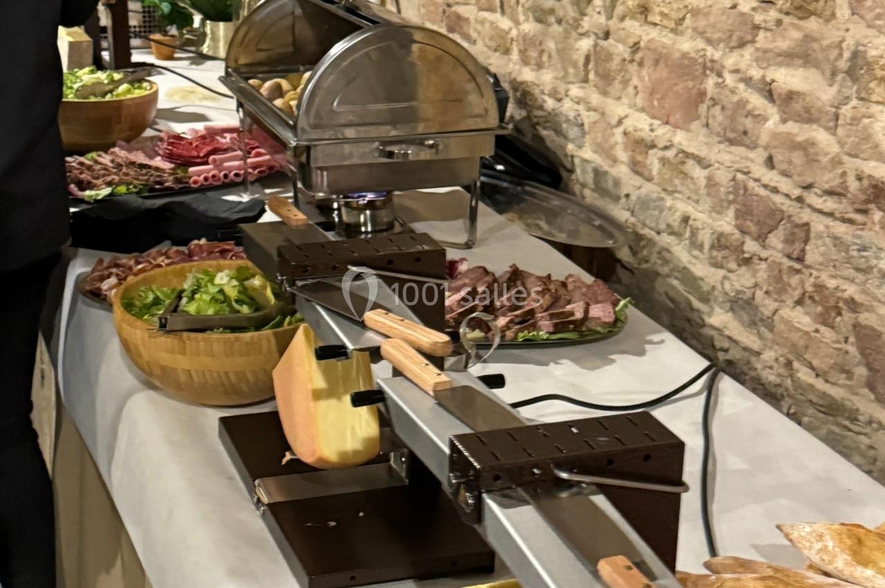 Table avec appareils à raclette, charcuterie, pain, légumes et accompagnements, dans un cadre en pierre rustique.