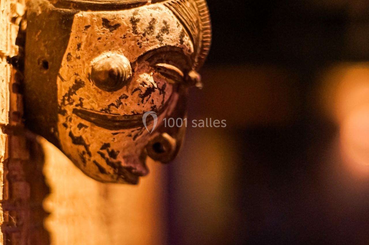 Masque sculpté en bois accroché à un mur, éclairé par une lumière chaude, avec un arrière-plan flou.