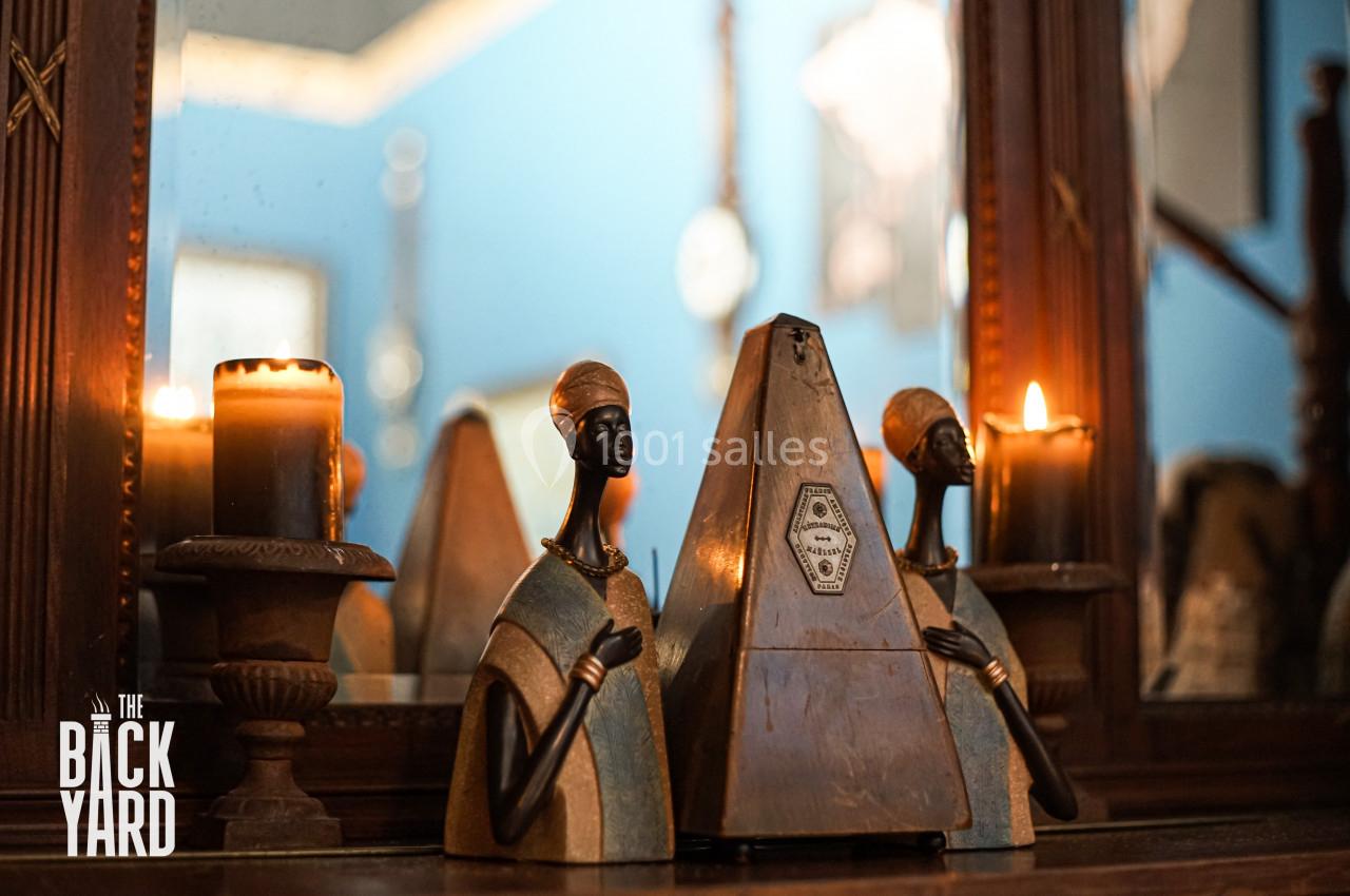 Deux statuettes africaines encadrent un métronome en bois, posés sur une table devant un miroir et des bougies allumées.