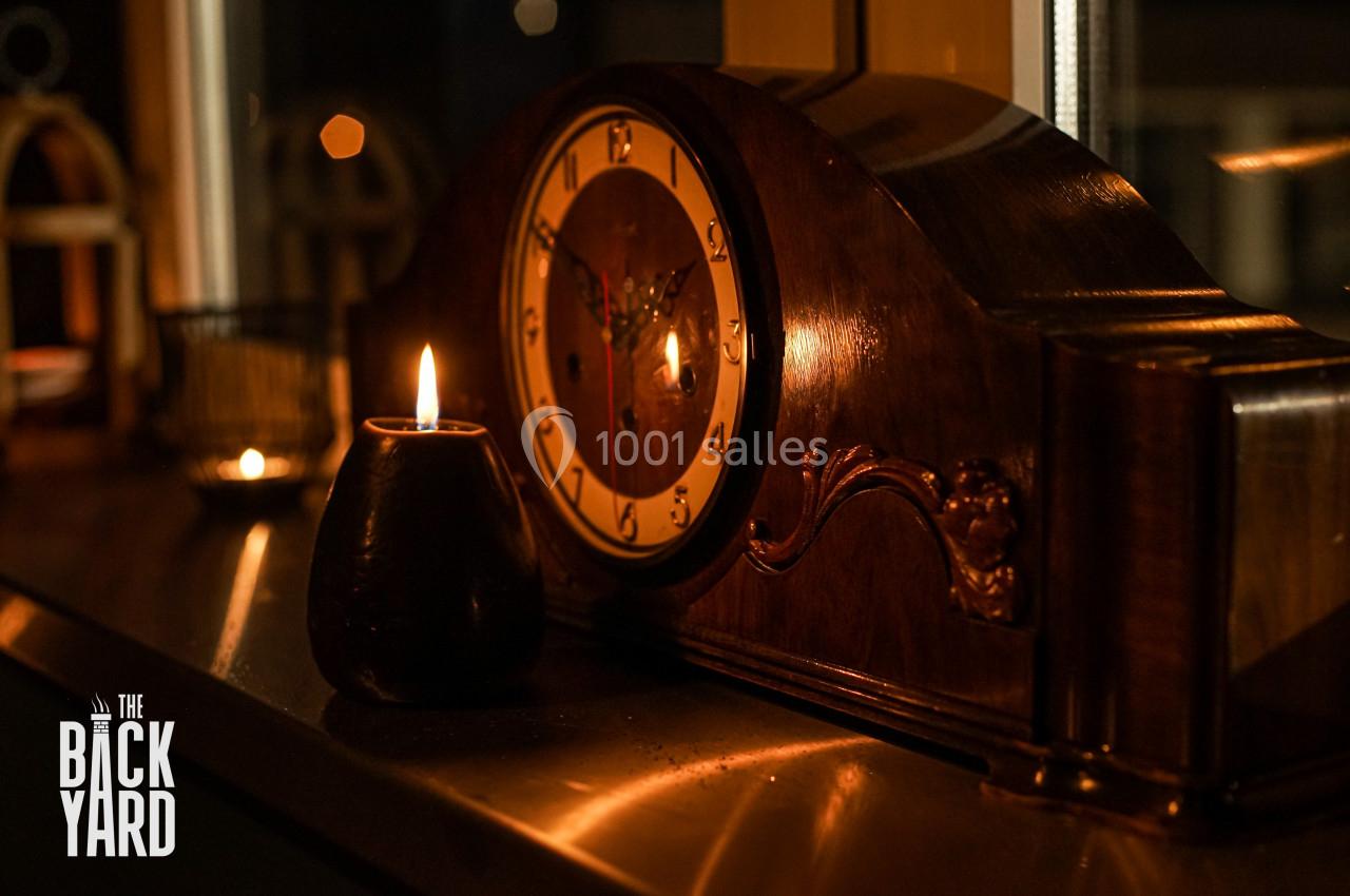 Horloge en bois rétro posée sur une surface, éclairée par une bougie allumée dans un environnement sombre.