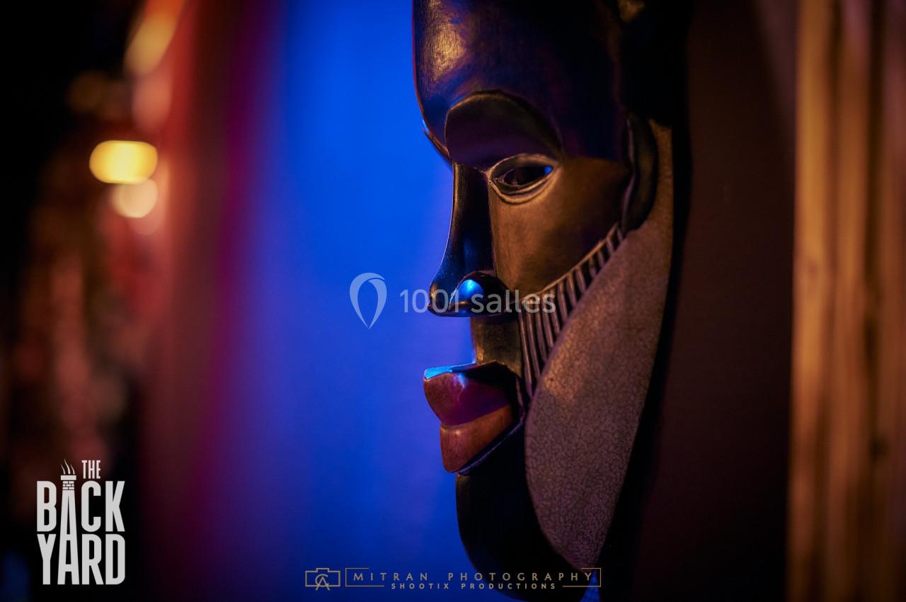 Masque sculpté en bois sombre accroché à un mur, éclairé par une lumière bleue et rouge dans un environnement tamisé.