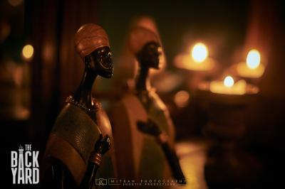 Deux statuettes africaines en gros plan, éclairées par des bougies en arrière-plan dans une ambiance tamisée.