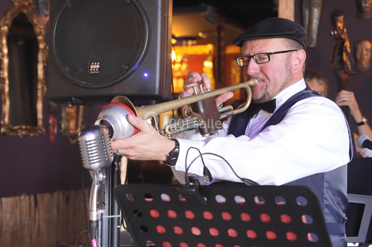 Un musicien joue de la trompette avec une sourdine dans un cadre intérieur décoré de masques et miroirs.