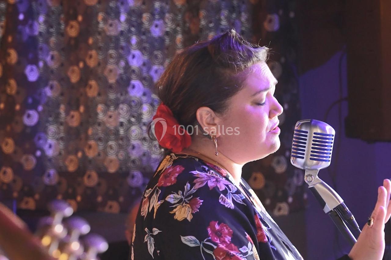 Une femme chante au micro dans une ambiance rétro, portant une robe à motifs floraux et une fleur rouge dans les cheveux.