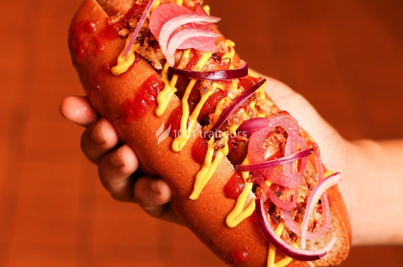 Hot-dog garni de moutarde, ketchup, oignons rouges et frits, tenu en main sur un fond flou orangé.