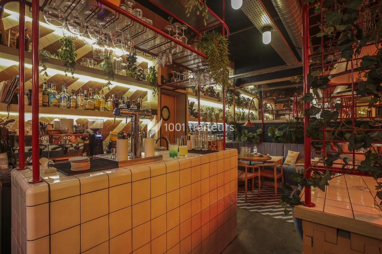 Intérieur d'un bar chaleureux avec comptoir carrelé, étagères garnies de bouteilles et plantes suspendues.