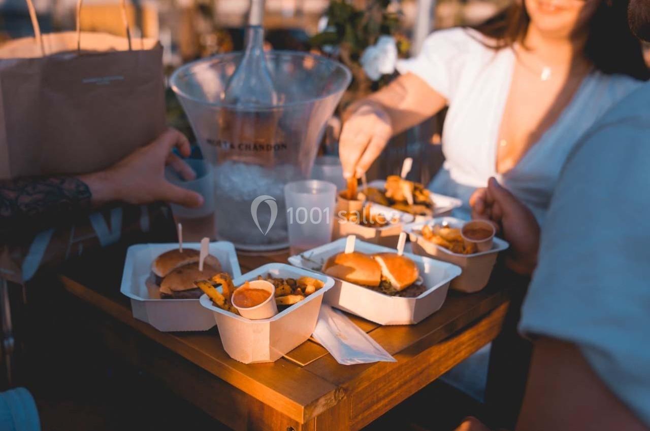 Repas en plein air avec des burgers, frites et sauces sur une table en bois, entourée de convives partageant le repas.